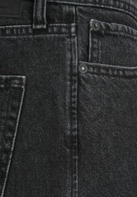 Tuvumā melnas džinsu bikses, kuras redzami jostas cilpas, priekšējie kabatas, šuvju detaļas un metāla kniedes.