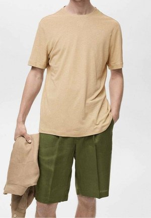 Mężczyzna w beżowej koszulce z krótkim rękawem i oliwkowo-zielonych szortach, trzymający beżową kurtkę w lewej ręce na jednolitym tle.