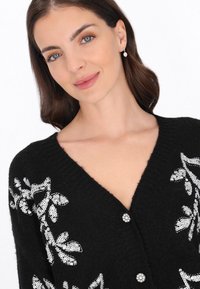 Fekete kardigán V-nyakkal, ezüst fényű flitteres virágmintával és két kerek, gyöngyszerű gombbal elöl. Strukturált kötött anyag.