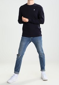 Person in einem navyblauen Langarmshirt, gerissenen blauen Jeans mit Aufschlägen, weißen Socken und weißen Sneakers, steht mit gespreizten Beinen und gefalteten Händen.