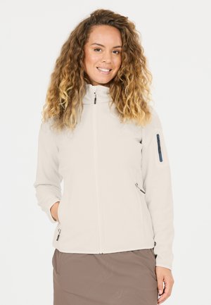 Kvinde med krøllet lyst brunt hår iført en lysebeige fleecetrøje med lynlås og brun nederdel, smilende med den ene hånd i jakke-lommen.