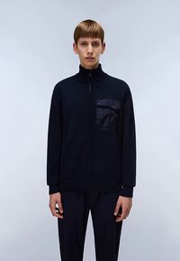 Marineblauw jack met ritssluiting, hoge kraag, geribbelde manchetten en een zak op de linkerborst met drukknopen; gedragen over bijpassende donkere broek.