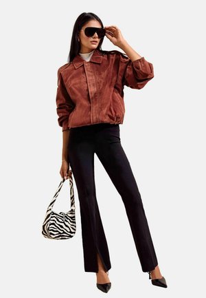 Frau trägt übergroße braune Jacke, schwarze ausgestellte Hose mit Schlitzen, schwarze spitze Absätze, große Sonnenbrille und hält eine Handtasche mit Zebramuster.