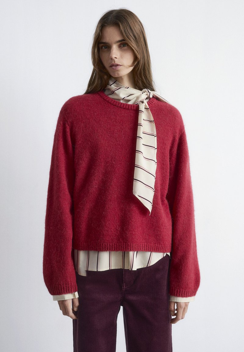 Red wool sweater with a cropped fit and ribbed cuffs, layered over a cream shirt with maroon vertical stripes, paired with dark corduroy pants.