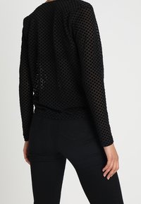 Top lungo a maniche nere in rete con ritagli circolari, scollatura rotonda e vestibilità ampia, abbinato a jeans skinny neri.