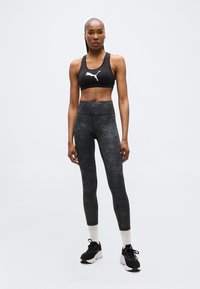 Αθλητική γυναίκα όρθια, φορώντας μαύρο αθλητικό σουτιέν Puma, σκούρο γκρι κολάν με ψηλή μέση, λευκές κάλτσες και μαύρα αθλητικά παπούτσια σε απλό φόντο.