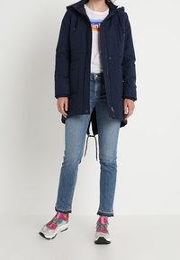 Granatowa pikowana parka z kapturem, dwoma przednimi kieszeniami i sznurkiem w pasie. W połączeniu z niebieskimi dżinsami i kolorowymi butami sportowymi.