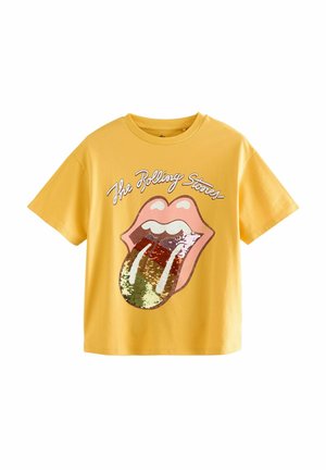 Žlté tričko s krátkym rukávom s logom Rolling Stones pier a jazyk zdobeným flitrami a bielym kurzívnym textom nad dizajnom.