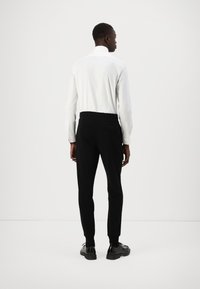 Weißes Langarmhemd mit runden Kragen, kombiniert mit schwarzen maßgeschneiderten Hosen. Glatter Stoff, schmaler Schnitt und schwarze Schuhe runden das Outfit ab.