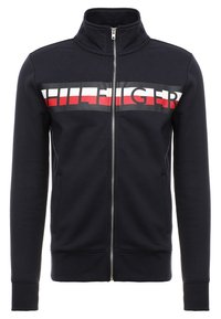Giacca con zip blu navy in misto cotone con collo alto, caratterizzata da un audace logo "HILFIGER" in rosso, bianco e nero sul petto, tasche laterali.