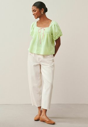 Woman wearing a light green puff-sleeve blouse with a pink tie, white high-waist cropped pants, and brown flats, standing with hands in pockets.