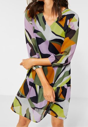 Kobieta w sukience z dekoltem w serek i długim rękawem, z kolorowym abstrakcyjnym wzorem w zielonym, fioletowym, pomarańczowym, czarnym i białym kolorze, stojąca na jednolitym tle.