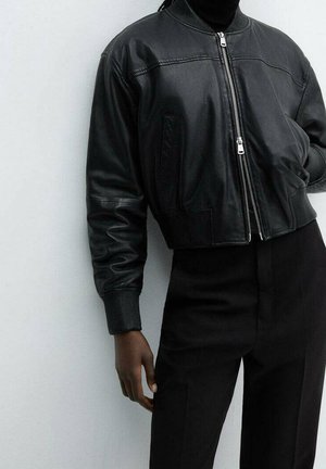 Personne portant une veste bomber en cuir noir sur un col roulé noir et un pantalon noir taille haute sur un fond gris clair.