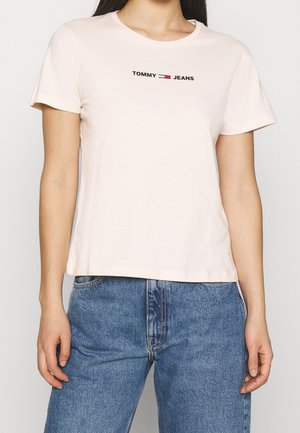 T-shirt en coton rose clair avec un col rond et des manches courtes, arborant un logo "TOMMY JEANS" noir et rouge centré sur la poitrine.