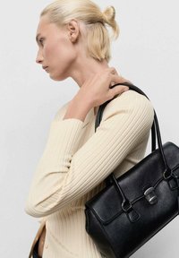 Borsa a tracolla in pelle nera con superficie liscia, accessori in argento, manico superiore e chiusura a patta, indossata sopra un maglione crema a coste leggero.