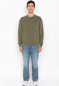 Olivgrüner Sweatshirt mit einem gerippten Kragen und Bündchen, kombiniert mit hellblauen Jeans mit geradem Bein und weißen Turnschuhen.