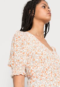 Vestido floral com fundo branco, apresentando flores laranja, decote em V e mangas curtas com mangas bufantes e punhos franzidos.