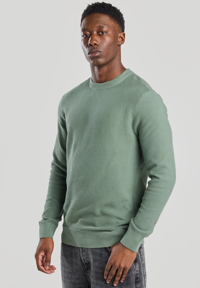 Green waffle-knit sweater features a round neck, long sleeves, and ribbed cuffs and hem. Paired with dark jeans, textured fabric detail visible.
