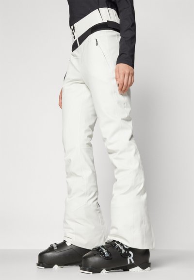 Witte skibroek met hoge taille, voorzien van een zwart accent en meerdere ritssluitingen. Draagt met zwarte skischoenen.