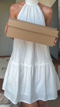 Bruine kartonnen doos vastgehouden met twee handen, rechthoekige vorm, gladde textuur, met een klep sluiting. Witte, verzamelde stoffen jurk met een uitlopende zoom.