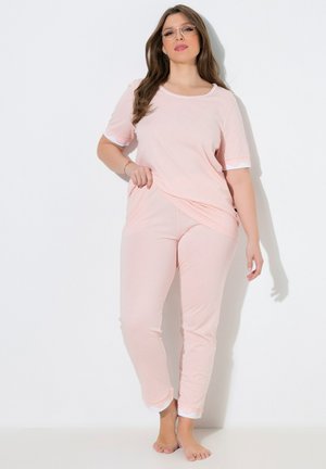 Mujer vistiendo un conjunto de ropa cómoda color rosa claro con ribetes blancos, de pie descalza contra una pared blanca, ajustándose la parte superior con una mano.