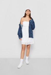 Hvit blond strapless kjole lagd med en denimjakke, komplimentert med hvite sneakers og en hvit caps. Glatt tekstur, uformell stil.