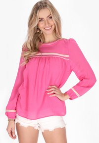 Blusa rosa con una finitura testurizzata, caratterizzata da un davanti arricciato, rifiniture dorate e maniche lunghe con polsini elastici, abbinata a pantaloni corti bianchi.