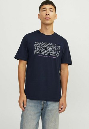 Mørkeblå bomulds t-shirt med hvide "ORIGINALS"-tekst i fed skrift. Klassisk rund hals og korte ærmer. Kombineret med lyseblå jeans.