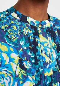Blouse florale avec un col mandarin, ornée de motifs floraux bleus, verts et jaunes sur un fond sombre. Plis et détails de boutons visibles.