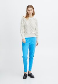 Helle, creamfarbene Strickpullover mit Rautenmuster, kombiniert mit blauen, figurbetonten Hosen und schwarzen Slippern, die eine glatte Textur und eine maßgeschneiderte Passform aufweisen.