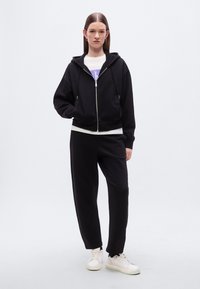 Μαύρο φούτερ με φερμουάρ και κορδόνια, συνδυασμένο με μαύρο παντελόνι jogger. Το άτομο φοράει λευκά αθλητικά παπούτσια, παρουσιάζοντας μια άνετη εφαρμογή και σχεδιασμό.