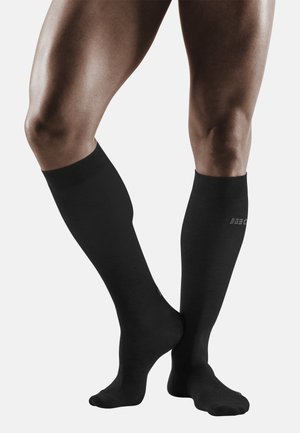 Schwarze knielange Socken aus elastischem Material, mit geripptem Bündchen und glatter Textur, auf einem Paar Beine platziert.