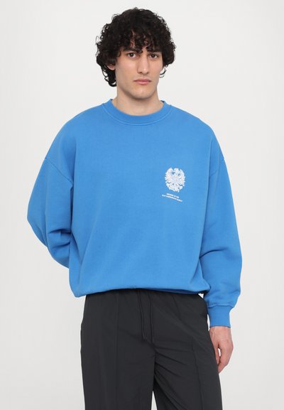 Blaues, oversized Sweatshirt aus weichem Stoff, mit einem weißen Emblem und Text auf der Brust sowie gerippten Bündchen und Kragen.