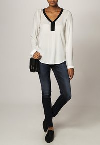 Blouse blanche à manches longues avec détail noir au col, associée à un jean skinny bleu foncé. Accessoires : petit sac à main noir et mocassins noirs.