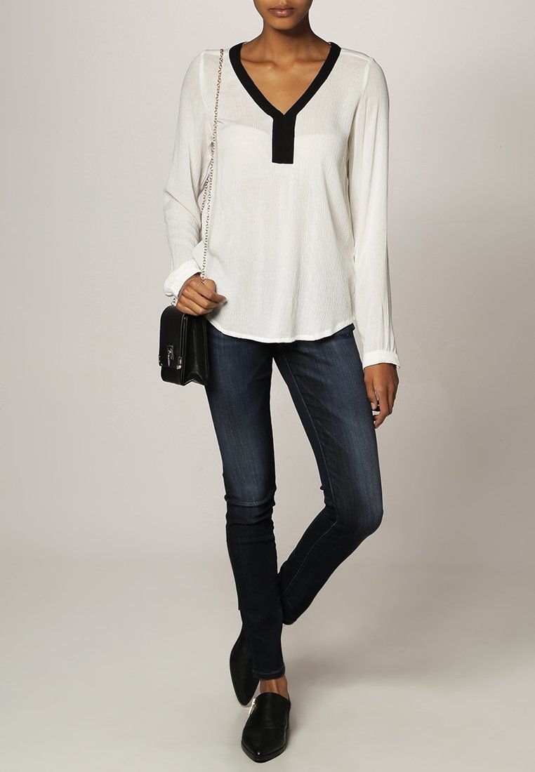 Blouse blanche à manches longues avec détail noir au col, associée à un jean skinny bleu foncé. Accessoires : petit sac à main noir et mocassins noirs.