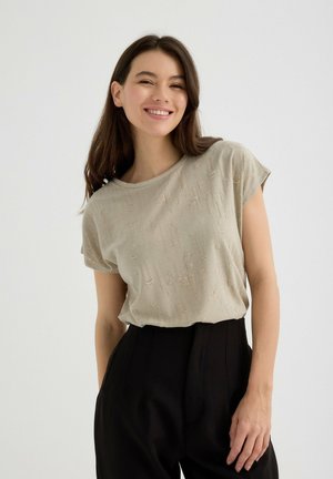 Camiseta de manga corta beige con un patrón sutil y texturizado. Diseño de cuello redondo combinado con pantalones negros de cintura alta.