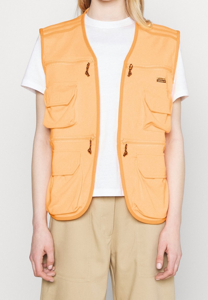 Orange tygväst med flera fickor, dragkedjeslutningar och orange detaljer. Bärs över en vit t-shirt och beige byxor.