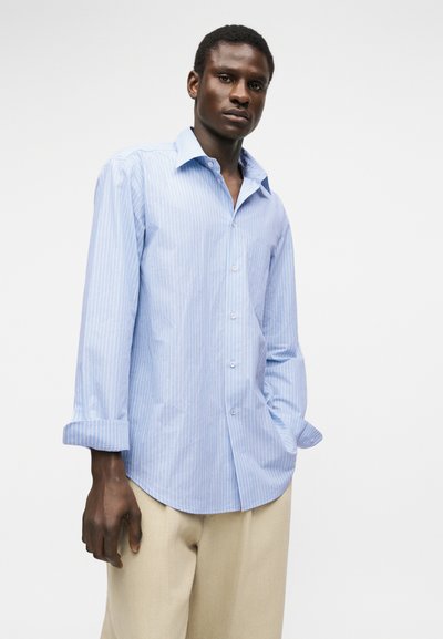 Άνδρας που φοράει ανοιχτό μπλε, μακρυμάνικο πουκάμισο με λεπτές λευκές ρίγες και μπεζ παντελόνι, στέκεται μπροστά σε απλό λευκό φόντο.