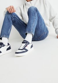 Zapatillas navy y blancas con cuero sintético texturizado, cierre con cordones y un logotipo de marca visible. Llevadas con jeans de color azul claro y calcetines blancos.
