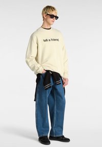 Sudadera clara con texto negro "dile a un amigo", combinada con jeans azules de gran tamaño y zapatillas negras. Los accesorios incluyen gafas de sol negras.