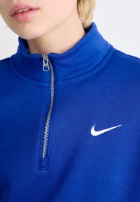 Modrý Nike pulovr s vysokým límcem, předním zipem na polovinu délky a bílým logem. Měkká tkanina s hladkým povrchem, navržená pro pohodlí.