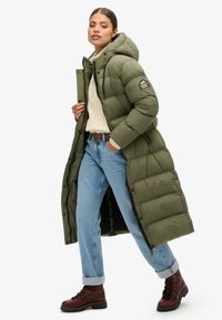 Manteau d'hiver vert olive long avec capuche, présentant une texture côtelée et un patch logo, associé à un jean bleu clair à revers et des bottes marron.