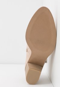 Botte beige à la cheville, vue en angle vers le haut montrant la semelle texturée et le talon bloc bas sur fond blanc.