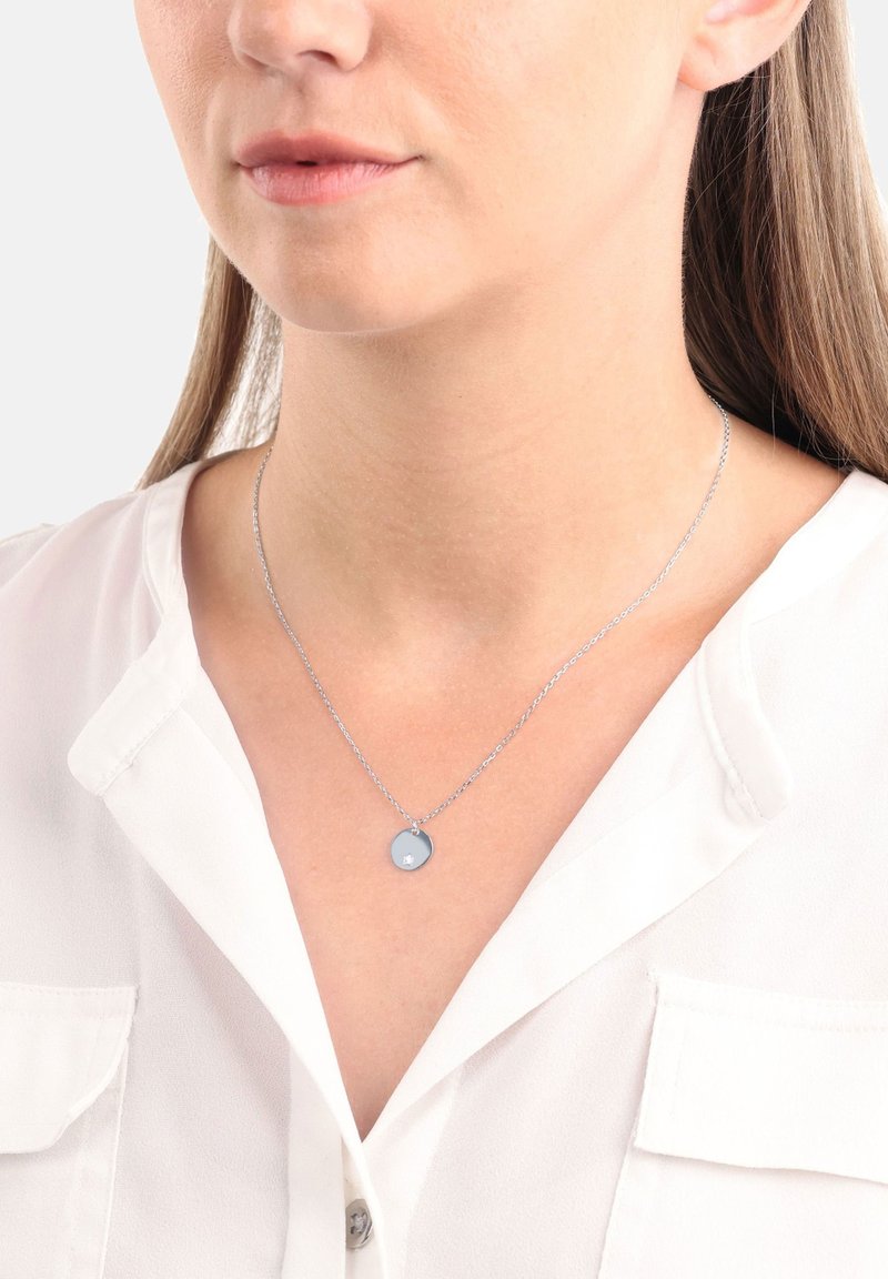 Silverhalsband med en cirkulär berlock, med en slät yta och en liten dekorativ detalj, bärs över en vit blus.