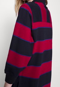 Maglietta a maniche lunghe in misto cotone con righe orizzontali rosse e blu navy con dettagli blu. Collo alto, vestibilità rilassata e maniche con polsini.