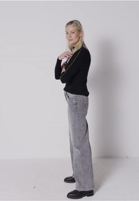 Czarny sweter z okrągłym dekoltem, szare szerokie spodnie dżinsowe i czarne botki za kostkę. Sweter ma kontrastowe wykończenia. Gładkie tło.