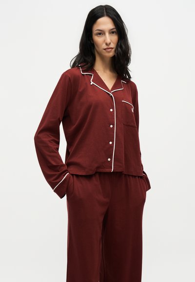 Femme aux cheveux noirs portant un pyjama boutonné rouge foncé avec un passepoil blanc, debout les mains dans les poches devant un fond uni.