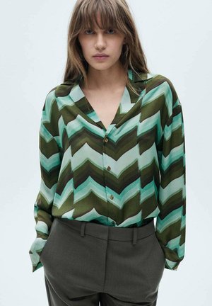 Bluza s chevron uzorkom u zelenoj i plavoj boji, s V-dekolteom i detaljima gumba, u kombinaciji s hlačama visokog struka u sivoj boji. Svilenkast materijal.