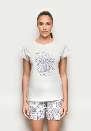 Frau trägt weißes T-Shirt mit maritimem Aufdruck und passende Shorts mit Muscheln, Seesternen und Seepferdchen vor einfarbigem Hintergrund.