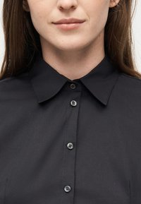 Γυναίκα που φοράει μαύρο πουκάμισο με κουμπιά και κλασικό γιακά, εμφανίζοντας το κάτω μισό του προσώπου και το πάνω μέρος του κορμού.